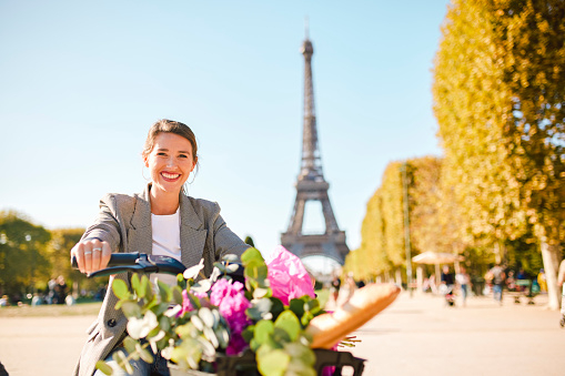 Female tourist smiling cheerfully at camera on electric bicycle with flowers, exploring park on sunny day, blurred view of Eiffel Tower in background