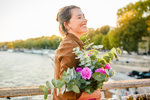 Happy young woman carrying flowers on bridge, smiling cheerfully with view of river in background