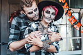 Scary love family couple man, woman celebrating halloween.Terrifying black skull half-face makeup,witch costumes,tattoo, stylish images,jacket,hat.Horror,fun at photoshoot, holiday party. Decorating of porch. Home, outdoor,street pumpkin jack-o-lantern