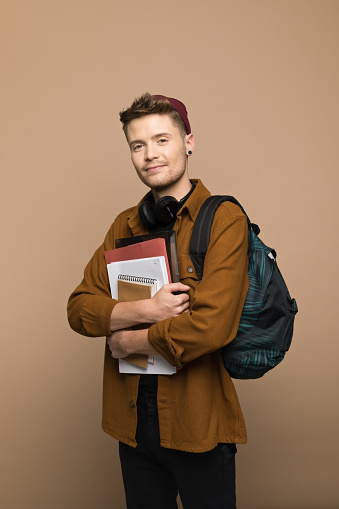 Portrait of confident young man wearing brown shirt, beanie and backpack, holding books and smiling at camera. Studio shot, grey background.