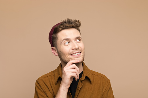 Headshot of thoughtful young man wearing brown shirt and beanie looking up with hand on chin. Studio shot, grey background.