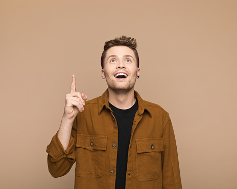 Portrait of happy young man wearing brown shirt and beanie pointing at copy space. Studio shot, grey background.