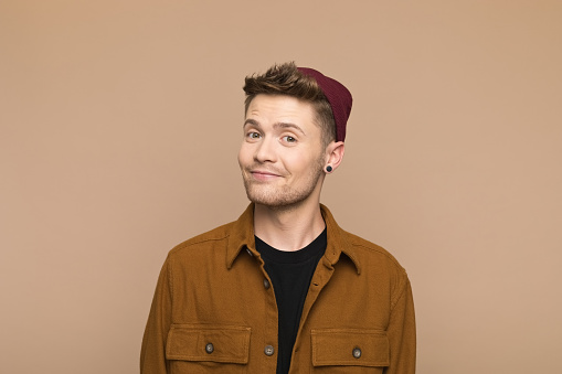 Portrait of happy young man wearing brown shirt and beanie smiling at camera. Studio shot, grey background.