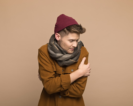 Portrait of young man wearing brown shirt, scarf and beanie hugging self. Studio shot, grey background.