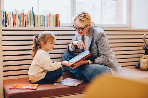 Beautiful woman wearing glasses and her young daughter sitting on the indoor bench in the library. They are smiling and bonding over reading books. Mother is holding the book while the girl is looking down and pointing at something.