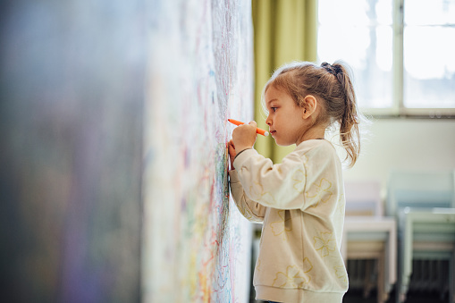 Smiling young girl painting with a felt tip pen on a big white piece of paper on a wall.