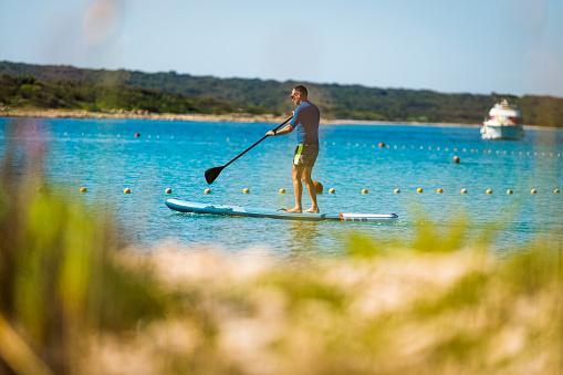 Man riding SUP stand up paddle on vacation. Active male riding SUP boards and paddling in the ocean on a sunny day. Athletic person on SUP board with yachts in background, Silba, Croatia.