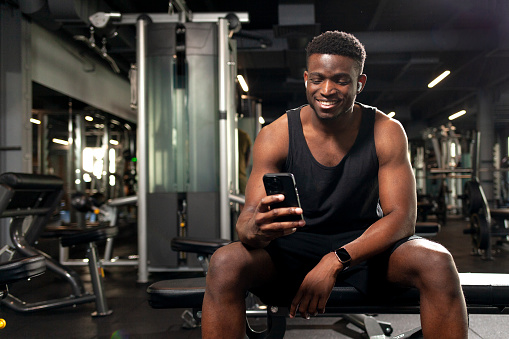 young athletic african american man sits in dark gym and uses smartphone, sports guy types online on the phone in fitness club and smiles