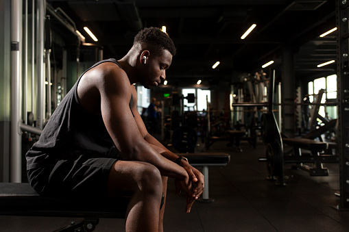 young athletic african american man sits in dark gym and thinks, pensive athletic guy rests and looks forward in fitness club, portrait of athlete in training