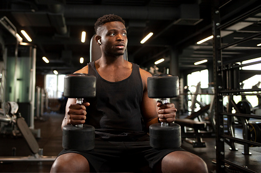 young athletic african american man trains in dark gym, athletic guy lifts heavy dumbbells in fitness club