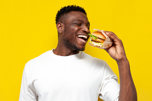 african american man in white t-shirt holding big burger and smiling, the guy eats fast food on yellow isolated background, the concept of unhealthy food and nutrition