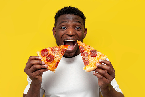 happy african american man in white t-shirt holding two slices of pizza and smiling over yellow isolated background, young funny guy eating fast food and screaming