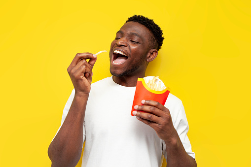 happy african american man in white t-shirt holding potato fries over yellow isolated background, young funny guy eating fast food and rejoicing