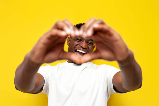 happy african american man in white t-shirt shows heart with his hands on yellow isolated background, toothy guy makes gesture of love and shows smile and face