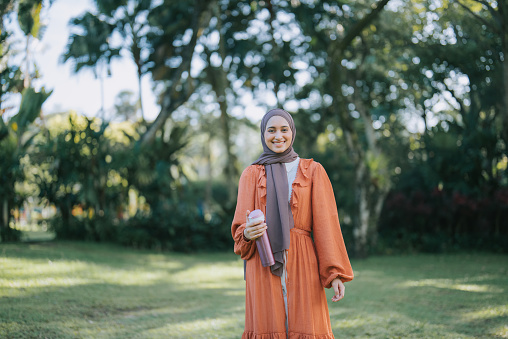 Asian Muslim young woman standing in public park smiling looking at camera with hijab