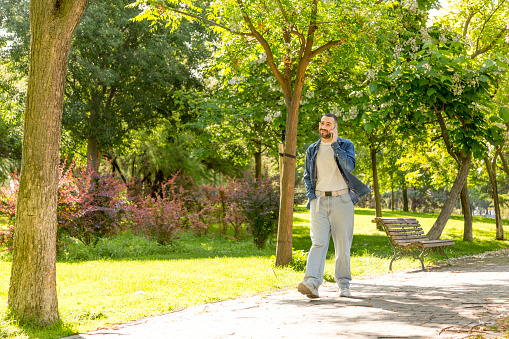 young bearded man, bathed in sunlight, strolling through a natural park while engaged in a phone conversation
