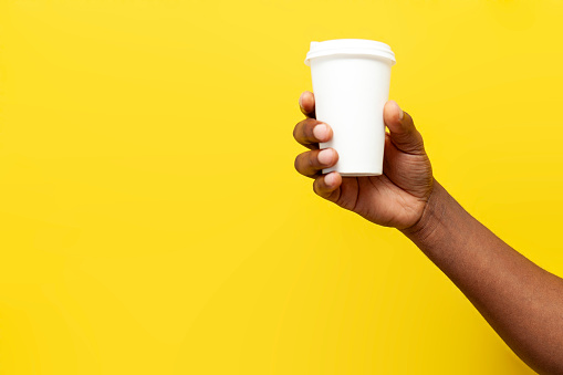 african american man's hand holds and offers white paper cup of coffee on yellow isolated background, empty package for drink in the hands of man, mock-up