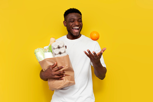 happy african american male shopper holding eco bag with groceries and nodding orange against yellow isolated background, guy customer rejoicing and smiling
