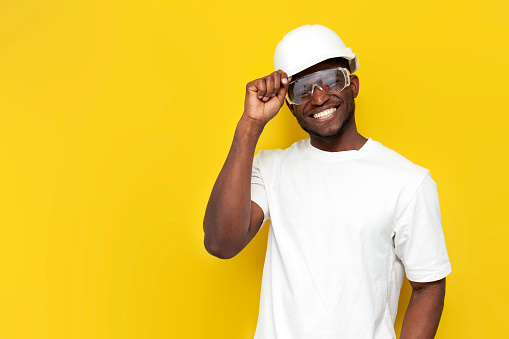 african american male civil engineer in hard hat and goggles smiles on yellow isolated background, builder in uniform stands on colored background