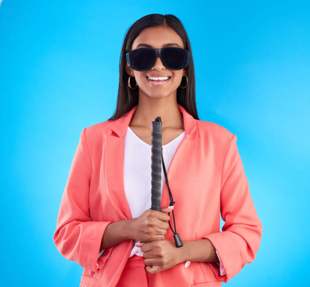 Blind, disability and walking stick with a woman on a blue background in studio wearing sunglasses. Portrait, vision and smile with an attractive young female holding her cane for disabled lifestyle