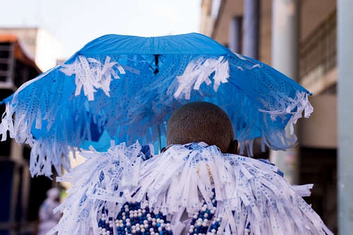 Salvador, Brazil – February 12, 2018: A person wearing a stylish and detailed ensemble is standing with their back to the camera, holding a blue umbrella in their hand