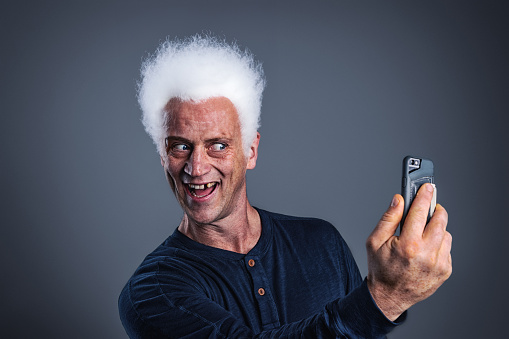 Portrait of an adult male with a missing tooth and funky white hairdo, taking a selfie with his smartphone.