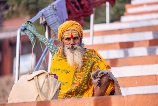 Varanasi, Uttar Pradesh, India - November 2022: Portrait of Unidentified Indian sadhu holy man in traditional saffron clothes sitting  in front of temple in varanasi city.