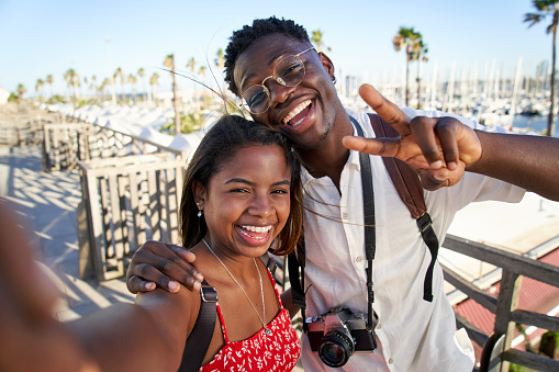 Happy young African tourist couple enjoying summer vacation in coastal resort town taking selfies while hanging out together.