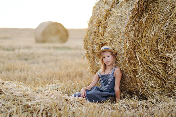 adorable caucasian girl enjoy having fun sitting neder golden hay bale on wheat harvested field near farm. happy childhood and freedom concept. rural countryside scenic landscape - wheat sunset bale autumn imagens e fotografias de stock