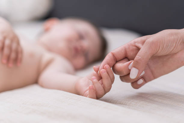 schläfriger kleiner junge greift mit winziger hand nach dem finger der mutter, die auf weichem bett mit weißem laken liegt - furniture barefoot little boys family stock-fotos und bilder