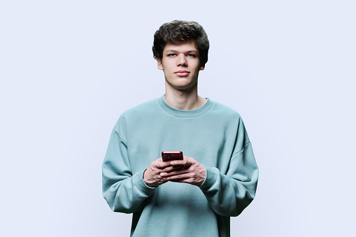 Young smiling guy college student with smartphone looking at camera on white background. Handsome teenage male with curly hairstyle. Internet technologies, mobile online applications for leisure learning services