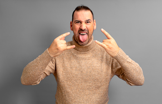 The show must go on. Photo of man sticking his tongue out of his mouth making horns sign rock and roll lover wearing beige turtleneck, isolated over gray background
