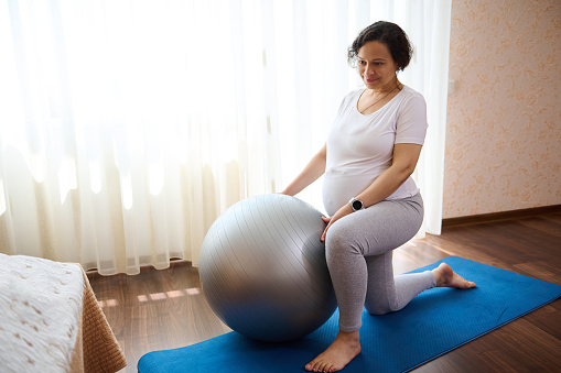 Confident pregnant woman exercising with fit ball, doing prenatal stretching and breathing exercises for healthy childbearing and easy delivery. Maternity concept. 9 month of active healthy pregnancy.