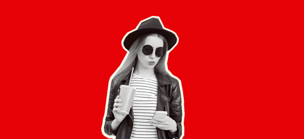 Portrait of stylish modern young woman with smartphone, cup of juice wearing black round hat, leather biker rock jacket on red background, magazine style stock photo