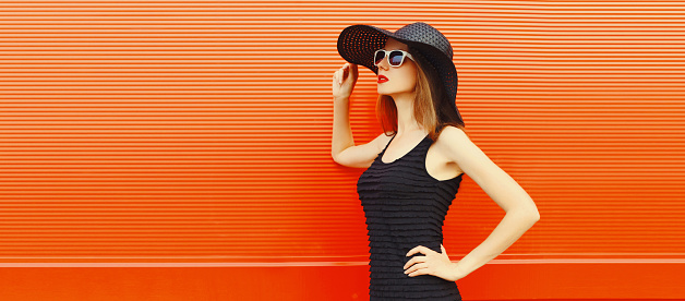 Portrait of beautiful young woman posing wearing black summer straw hat and dress looking away on orange background, blank copy space for advertising text