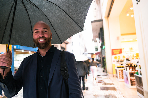 Horizontal medium shot of stylish Caucasian man wearing eyeglasses standing outdoors under transparent umbrella holding coffee cup, looking away and smiling