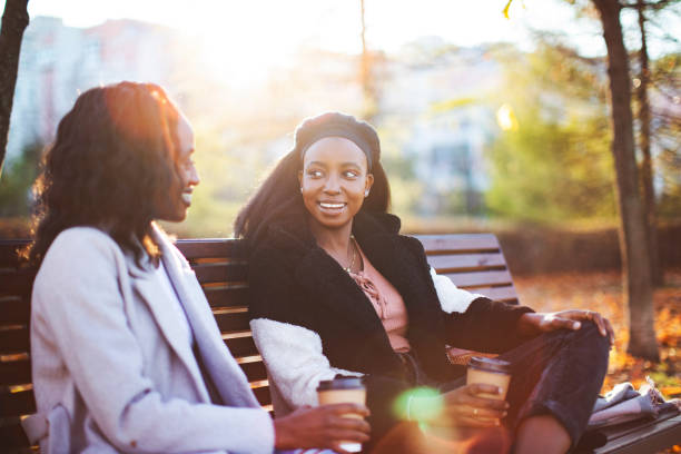 istock Two young black female friends sitting on park bench, drinking coffee and talking 1498220511
