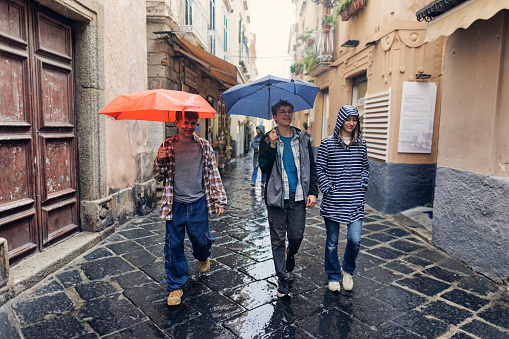 Teenagers walking in the rain in an Italian town. They are wearing raincoat and carrying umbrellas.\nCanon R5