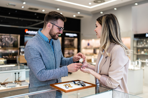 Elegant middle age businessman choosing and buying his new expensive watch. Beautiful youg female seller helps him to make good decision. Fashion style and elegance concept.