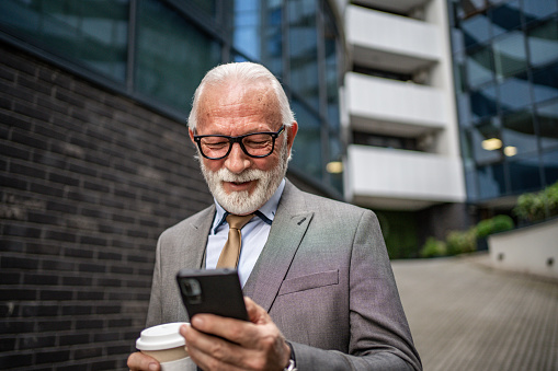 A shot of a mature businessman on the go using his smartphone and drinking takeaway coffee, confidence and vast business experience showing on his face and attitude.