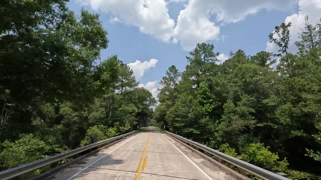 Driver view across small concrete bridge over forest creek with clouds forming in the sky
