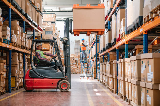 uniformed worker driving and loading cardboard boxes with forklift stacker loader in a factory warehouse - lagerlokal bildbanksfoton och bilder