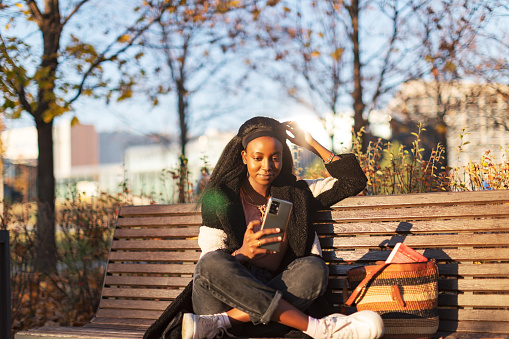 Young adult black woman sitting on park bench and using mobile