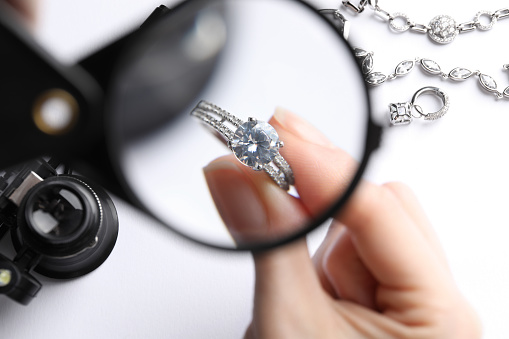 Jeweler examining diamond ring with magnifying glass at white table, closeup