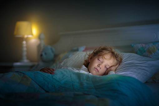 Child sleeping in dark bedroom. Little boy napping. Healthy night rest. Kids room with night light. Toddler with teddy bear. Bedding for children.