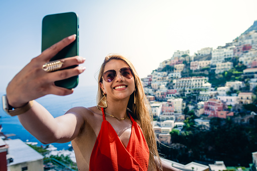 beautiful latin woman taking a selfie, with a nice red dress, in the background you can see an Italian coast, houses and a summer sun and sea.