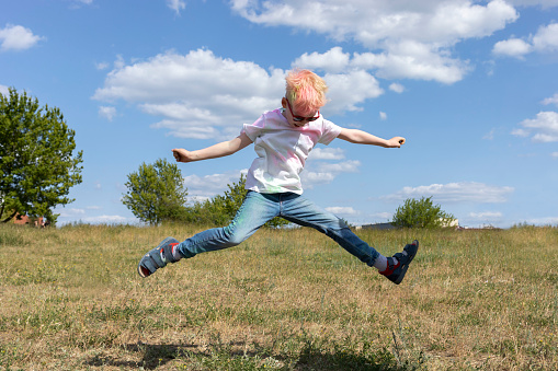 Little Blonde Boy With Colorful Paint, Powder On Clothes Jump Up in Air On Holi Color Festival. Emotional Happy Child. Blue Sky, Green Trees On Background, Summer. Horizontal Plane. High quality photo
