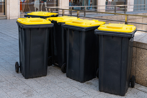 A group of black plastic trash cans with yellow lids stands in the pedestrian part of the city..