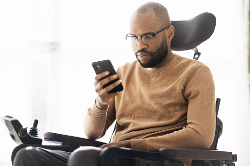 Side view of bearded black male in casual clothes and eyeglasses sitting on wheelchair in living room and browsing smartphone
