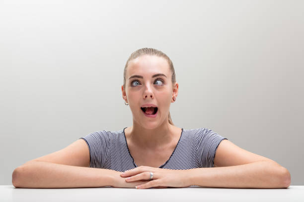 Young woman rests forearms on table, crosses eyes and contorts face in jest. Strives for ridiculous and ugly, but her beauty only evokes smiles and amusement